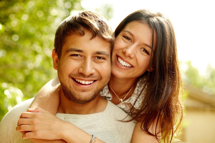 a woman hugs a man from behind while smiling at the camera